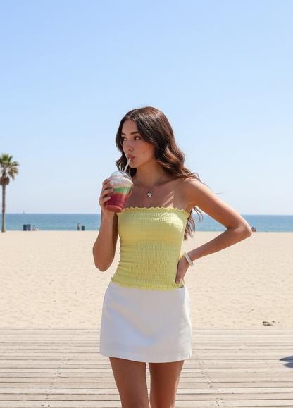 Model wearing an ÁMBER Del Mar lettuce trim yellow smocked tube top on a sunny beach, ideal for a coastal-chic summer outfit