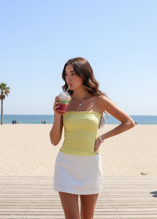Model wearing an ÁMBER Del Mar lettuce trim yellow smocked tube top on a sunny beach, ideal for a coastal-chic summer outfit