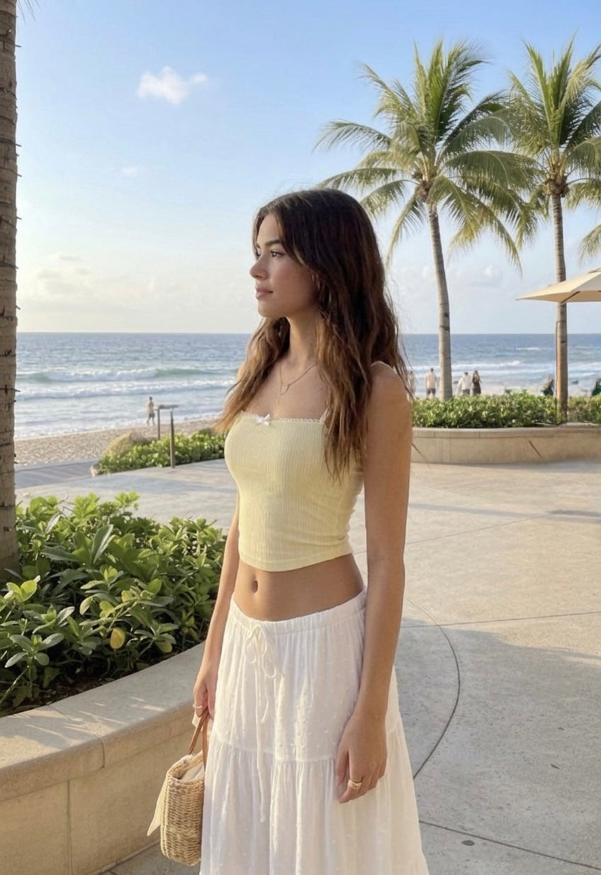 A young woman posing on a beach with a kissy face expression, wearing a yellow tank top and white skirt from ÁMBER Del Mar, embodying the clean girl aesthetic