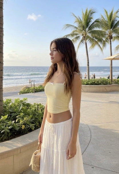 A young woman posing on a beach with a kissy face expression, wearing a yellow tank top and white skirt from ÁMBER Del Mar, embodying the clean girl aesthetic