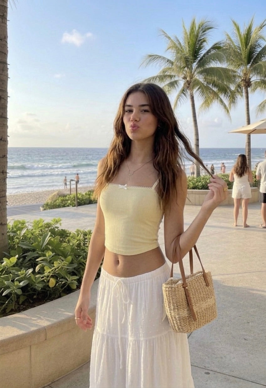 A young woman posing on a beach with a kissy face expression, wearing a yellow tank top and white skirt from ÁMBER Del Mar, embodying the clean girl aesthetic