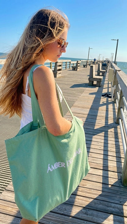 A model on a wooden pier carrying an oversized seafoam green canvas tote bag by Amber Del Mar.