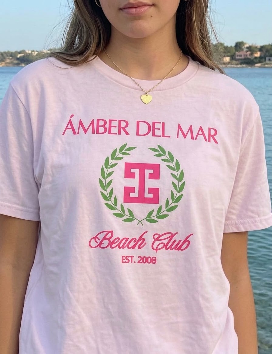 A teen girl wearing a light pink graphic t-shirt with 'ÁMBER DEL MAR Beach Club EST. 2008' printed in pink and a green laurel wreath logo, set against a background of the ocean.
