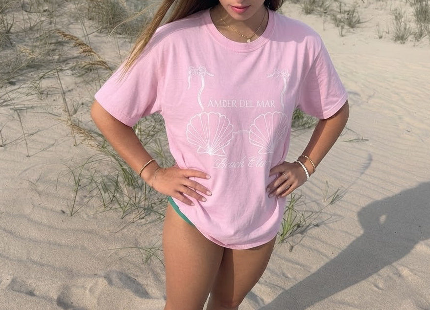 A teen girl standing on a sandy beach wearing a light pink graphic t-shirt with a seashell branded by Amber Del Mar.