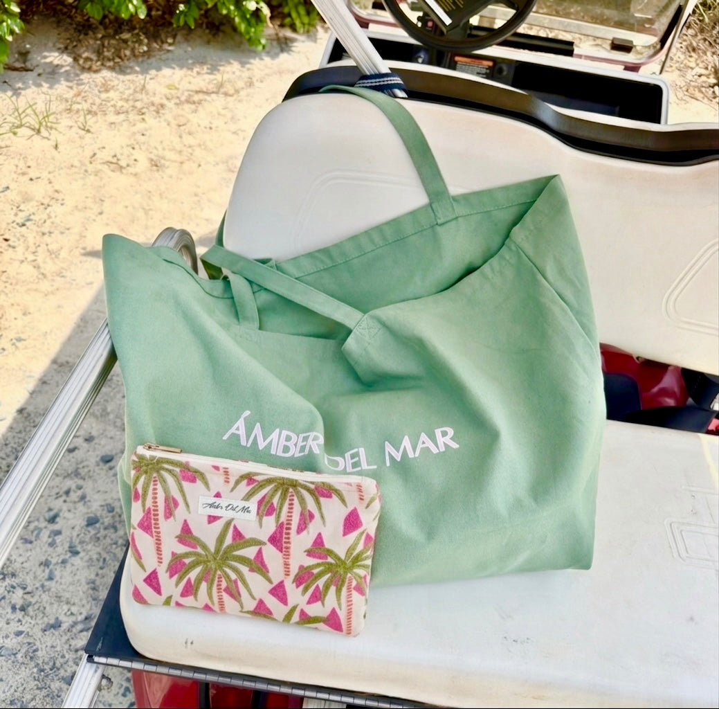 A seafoam green canvas tote bag by AMBER DEL MAR text sits on a white golf cart seat outdoors, next to a small green and pink palm tree patterned clutch.