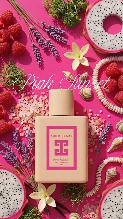 A perfume bottle labeled 'Amber Del Mar' and 'Pink Sunset' is centered on a bright pink background surrounded by decorative props including raspberries, dragon fruit slices, lavender, flowers, and seashells. The words 'Pink Sunset' appear as a large script overlay across the top.