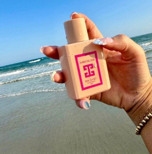 A hand with beachy nails holding a pink 50ml bottle of Amber Del Mar Pink Sunset Eau de Parfum against a beach background with ocean waves and a blue sky.