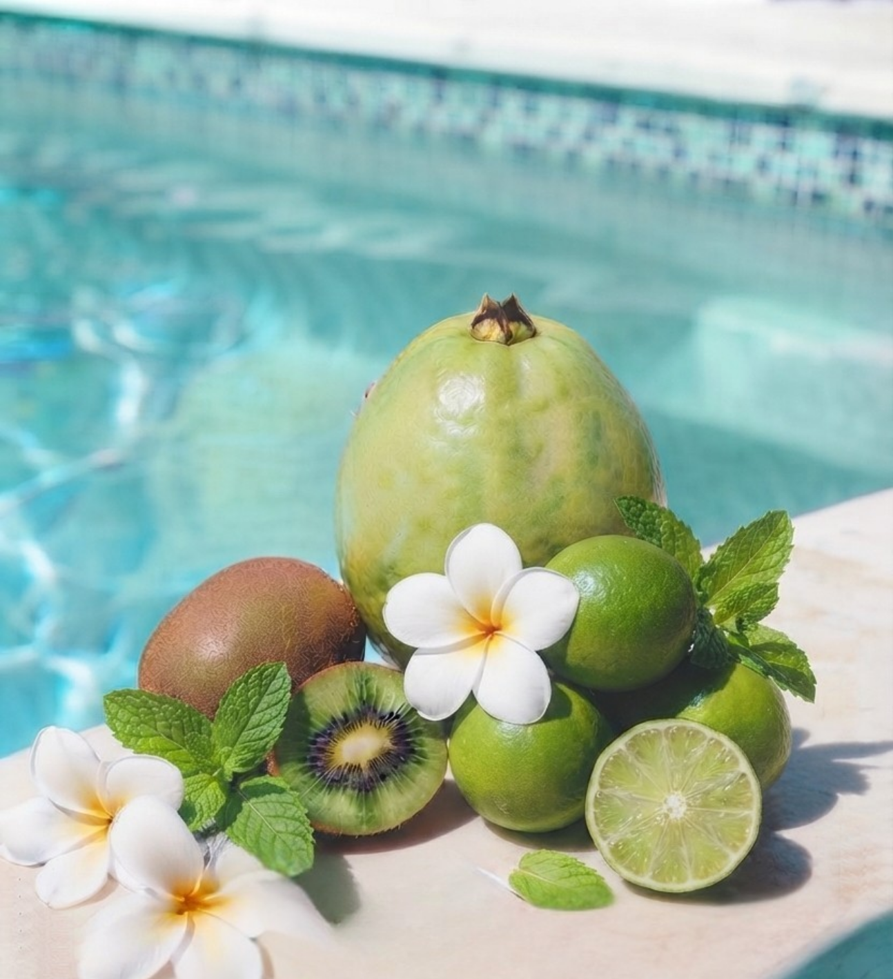 Flat lay composition of ingredients for ÁMBER DEL MAR Pretty Girl fragrance: whole mango, halved kiwi, and fresh limes and mint beside white gardenia flowers on a poolside ledge.
