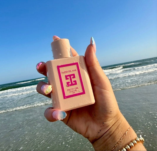 A hand with beachy nails holding a pink 50ml bottle of Amber Del Mar Pretty Girl Eau de Parfum against a beach background with ocean waves and a blue sky.