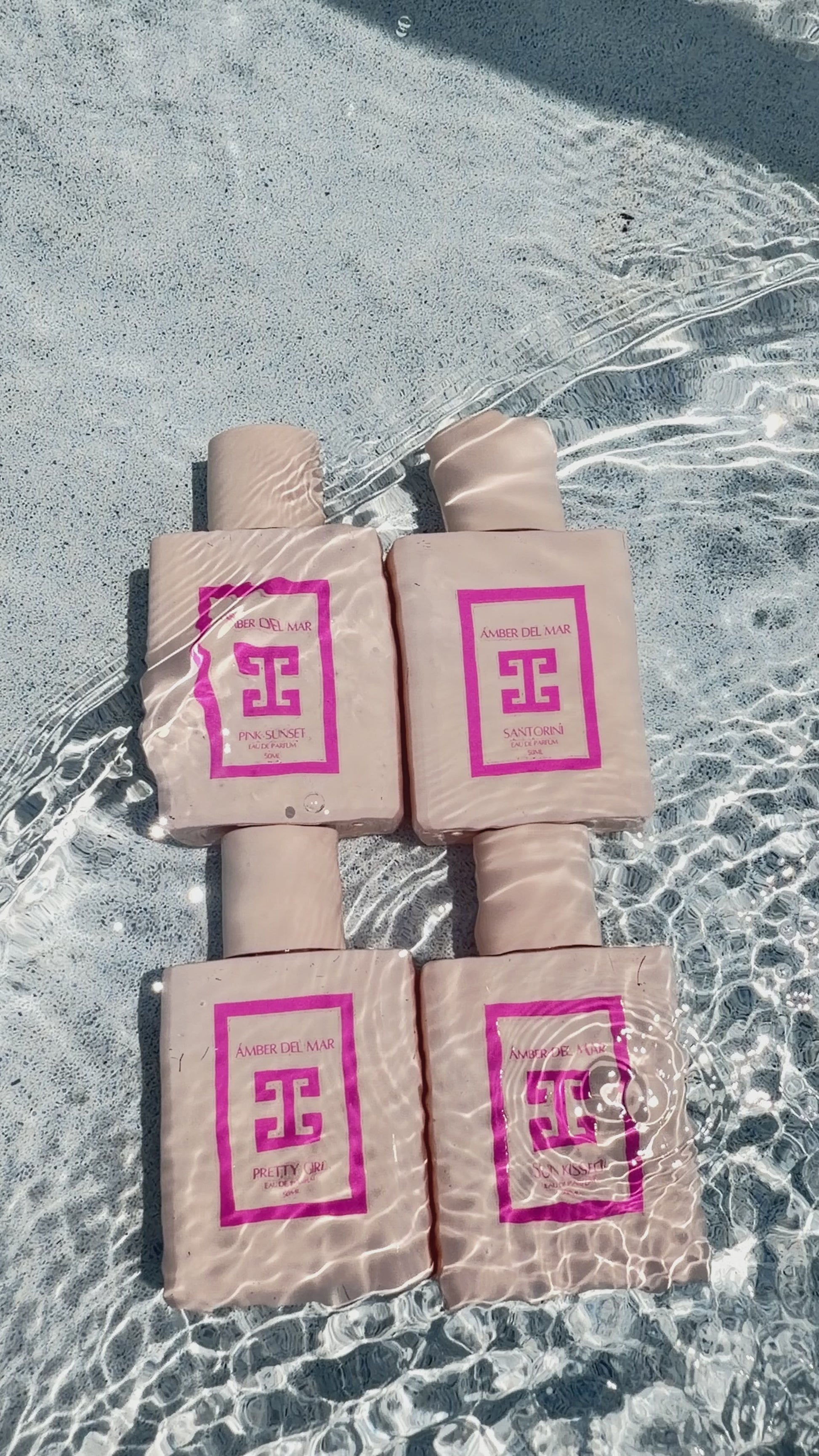 A video showing four AMBER DEL MAR perfume bottles in crystal clear water. Water continuously flows over them, with the sunlight hitting them and highlighting a minimalist pink bottles.