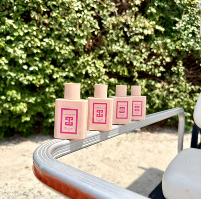 Four pinkperfume bottles by Amber Del Mar lined up on the curved silver arm of a golf cart, with a green hedge and sandy ground in the background.