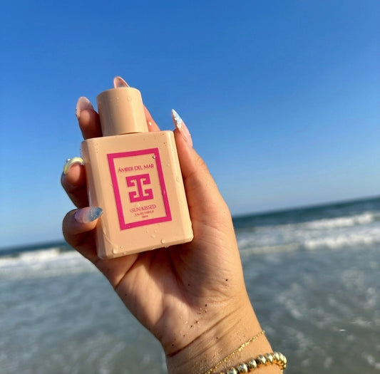 A hand with beachy nails holding a pink 50ml bottle of Amber Del Mar Sunkissed Eau de Parfum against a beach background with ocean waves and a blue sky.