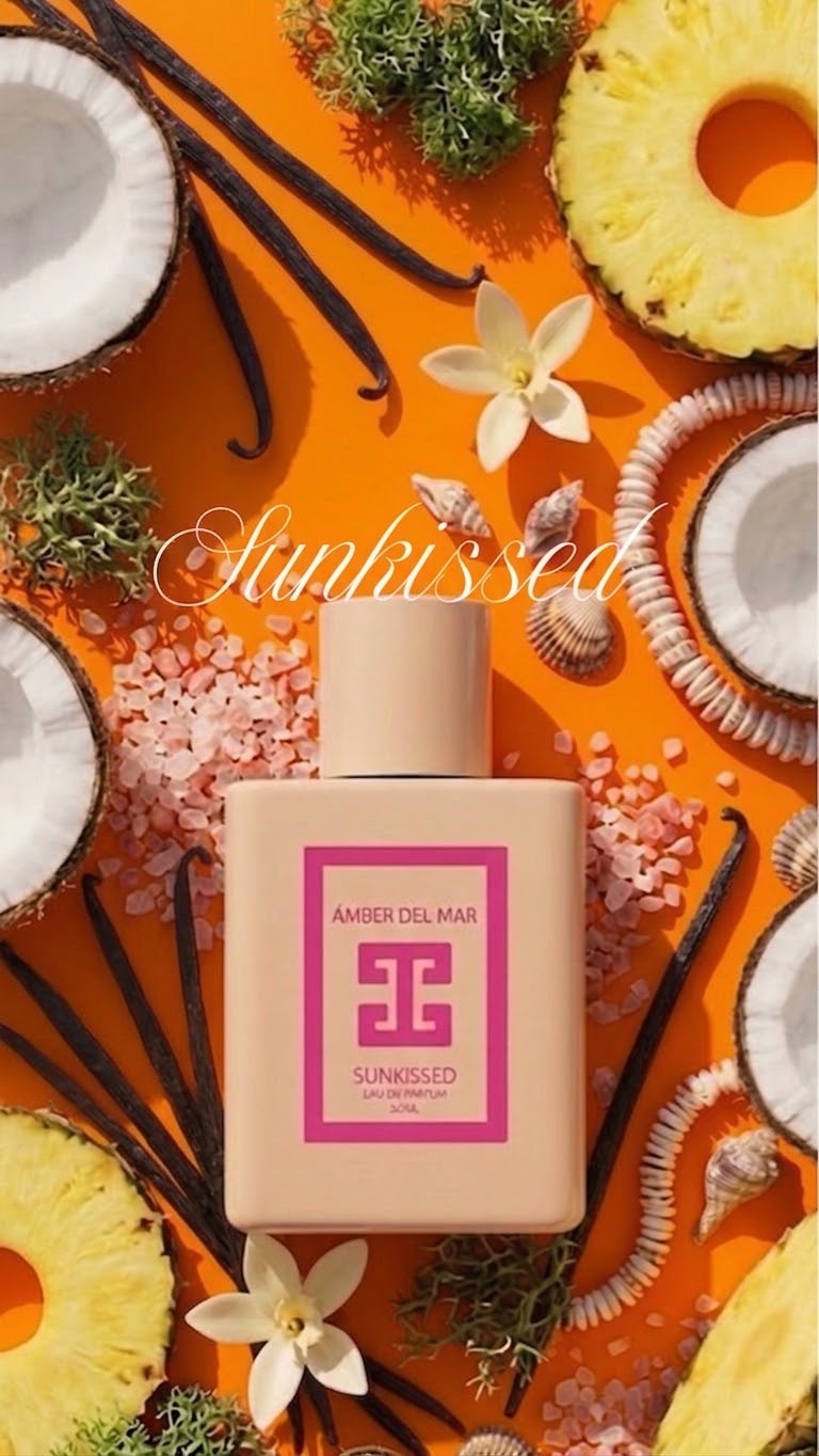 Amber Del Mar Sunkissed perfume bottle in a pink color with a pink label, styled in a flat-lay composition on an orange background with pineapple, coconut, vanilla beans, and flowers.